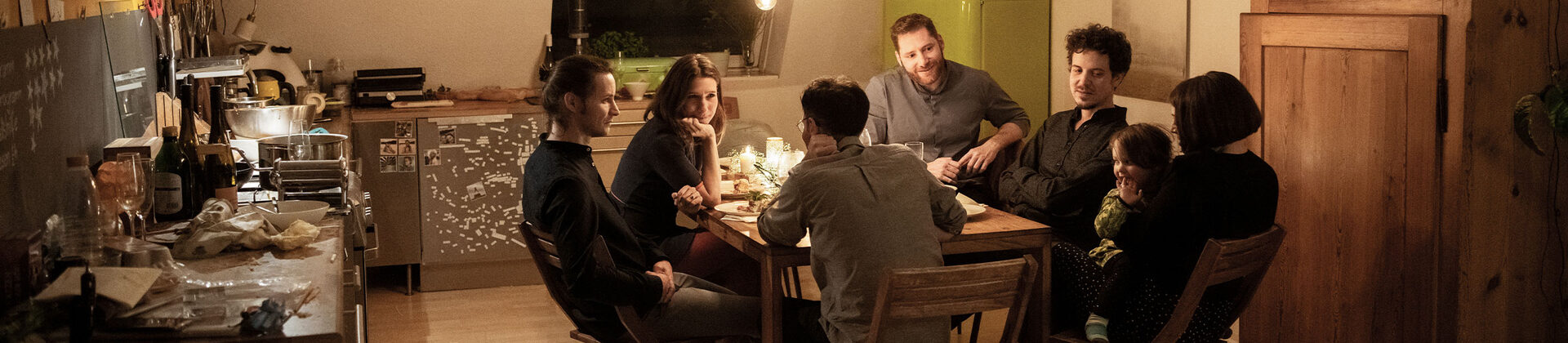 In einer Küche sitzt an einem Tisch eine Gruppe Menschen zusammen und unterhält sich.