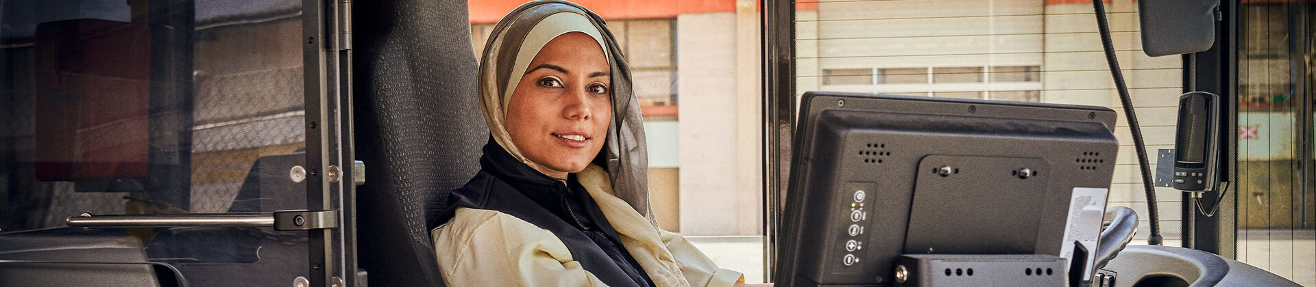Eine junge Frau mit Kopftuch sitzt am Steuer eines Busses und lächelt in die Kamera