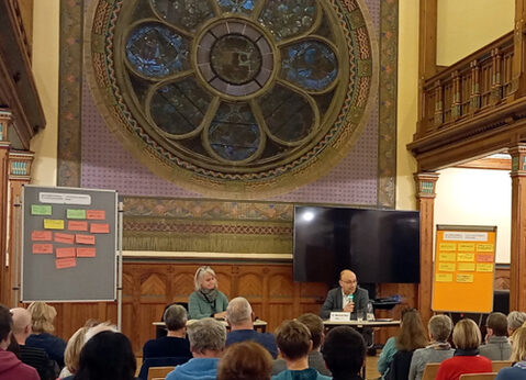 Ein Veranstaltungsraum mit hoher Decke und großem, kunstvoll gestaltetem Rundfenster im Hintergrund. Zwei Personen sitzen an einem Tisch auf einer Bühne und sprechen zu einem Publikum. Links und rechts der Bühne sind Pinnwände mit farbigen Notizzetteln aufgestellt. Die Zuhörerinnen und Zuhörer sitzen auf Stühlen und folgen der Präsentation