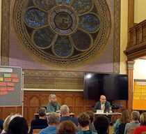 Ein Veranstaltungsraum mit hoher Decke und großem, kunstvoll gestaltetem Rundfenster im Hintergrund. Zwei Personen sitzen an einem Tisch auf einer Bühne und sprechen zu einem Publikum. Links und rechts der Bühne sind Pinnwände mit farbigen Notizzetteln aufgestellt. Die Zuhörerinnen und Zuhörer sitzen auf Stühlen und folgen der Präsentation