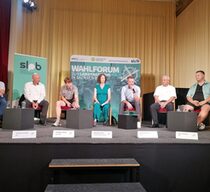 Sieben Personen sitzen auf einer Bühne, auf der sich im Hintergrund ein Banner mit der Beschriftung "Wahlforum zur Landtagswahl in Sachsen 2024" befindet. Von links nach rechts sind die Teilnehmer in einer Reihe auf Stühlen platziert. Vor jedem Teilnehmer steht ein Namensschild. Eine Person spricht in ein Mikrophon und die anderen hören zu.