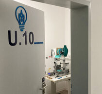 In einem Flur mit weißen Wänden befindet sich eine Tür mit der Aufschrift „U.10 Workspace“, die teilweise geöffnet ist und einen Raum mit einem 3D-Drucker und einem Tisch mit verschiedenen Objekten offenbart. Auf der rechten Seite hängt eine blaue Pinnwand mit Postern und Informationen.