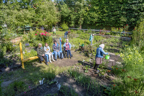 In einem großen, grünen Garten sitzen einige leute auf einer Bank und unterhalten sich, ander gießen Pflanzen oder erledigen andere Gartenarbeiten.