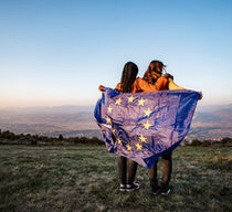 Zwei junge Frauen stehen Arm in Arm auf einer Wiese und gucken in die Ferne über Berge. Um ihre Schultern haben Sie eine Europa-Fahne gelegt.