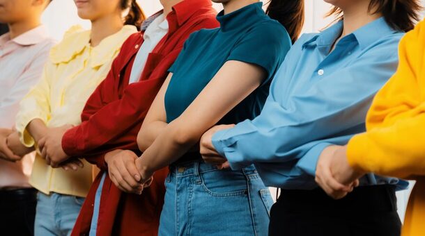 Personen unterschiedlichen Geschlechts in farbenfrohen Outfits stehen in einer Reihe zusammen mit überkreuzten Armen und gehaltenen Händen