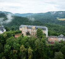 Ansicht der Schloss Schwarzburg in Thüringen aus der Luftperspektive. Das Schloss liegt auf einer Anhöhe und ist von einem Wald umgeben.