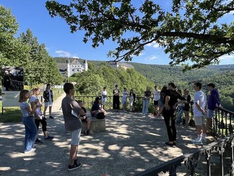 Aufnahme während der Projektumsetzung auf Schloss Schwarzburg im Jahr 2024. Die Teilnehmenden stehen im Kreis und lauschen einem Vortrag. Im Hintergrund sieht man zwischen Bäumen die Gebäude der Schwarzburg.