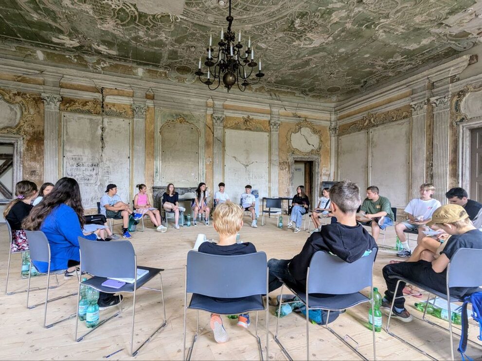 Die jungen Teilnehmenden sitzen in einem großen Stuhlkreis in einem Saal im Schloss Schwarzburg und unterhalten sich.