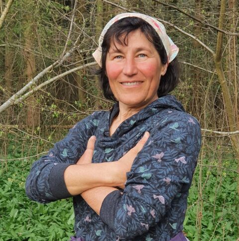 Eine Frau mittleren Alters mit schulterlangen, dunklen Haaren, die freundlich lacht. Ihre Arme sind vor dem Körper verkreuzt.