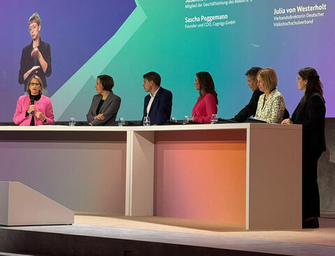 Sieben Personen stehen auf einem Podium, eine Frau in pinkem Jackett spricht in gestikulierend in ein Mikrofon.