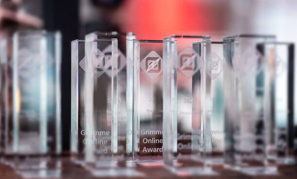 Viele Grimme Online Awards stehen auf einem Tisch. Das türkisblaue und rote Umgebungslicht bricht sich in den gläsernen Trophäen.