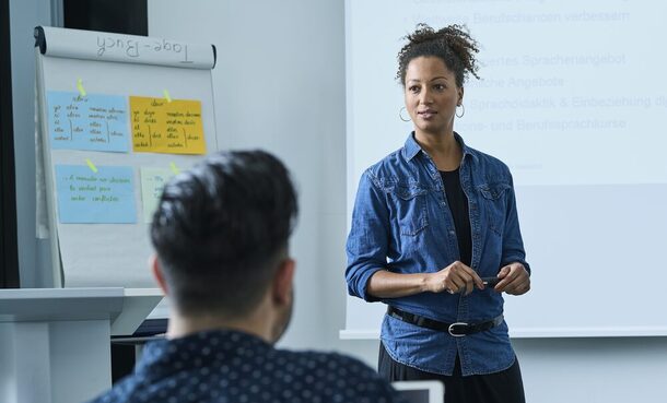 Eine junge Frau in einem Jeanshemd steht vor einer Klasse und unterrichtet