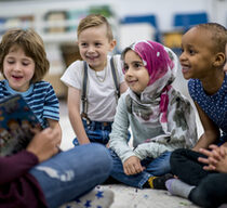 Eine multiethnische Gruppe von Schulkindern sitzt in einem Klassenzimmer. Sie tragen legere Kleidung. Sie sitzen auf dem Boden und hören ihrer Lehrerin gespannt zu, die ein Bilderbuch vorliest.