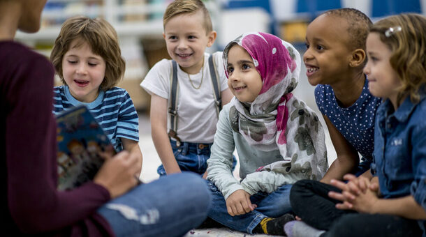 Eine multiethnische Gruppe von Schulkindern sitzt in einem Klassenzimmer. Sie tragen legere Kleidung. Sie sitzen auf dem Boden und hören ihrer Lehrerin gespannt zu, die ein Bilderbuch vorliest.