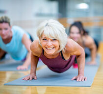 Eine Frau im mittleren Alter macht in einem Sportkurs auf dem Boden eine Plank und lächelt in die Kamera