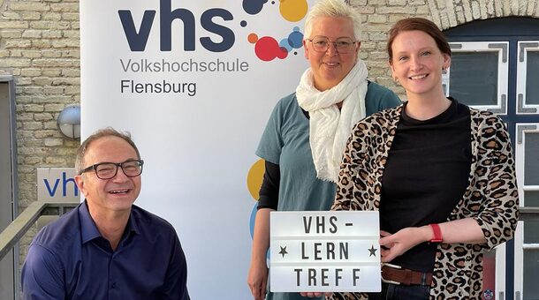 Drei Menschen stehen vor Schild der Volkshochschule Flensburg.
